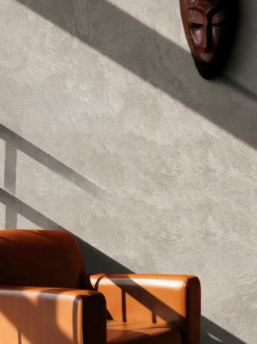 Wand met fijne metallic damasceringtextuur in scherp strijklicht, houten Afrikaans masker en cognac leren fauteuil